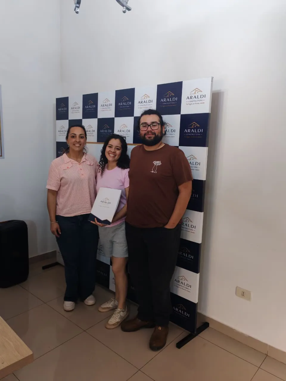 Tami, Junior y Yami Leiva de pie frente al fondo de Araldi. Tami sostiene la carpeta de la constructora.