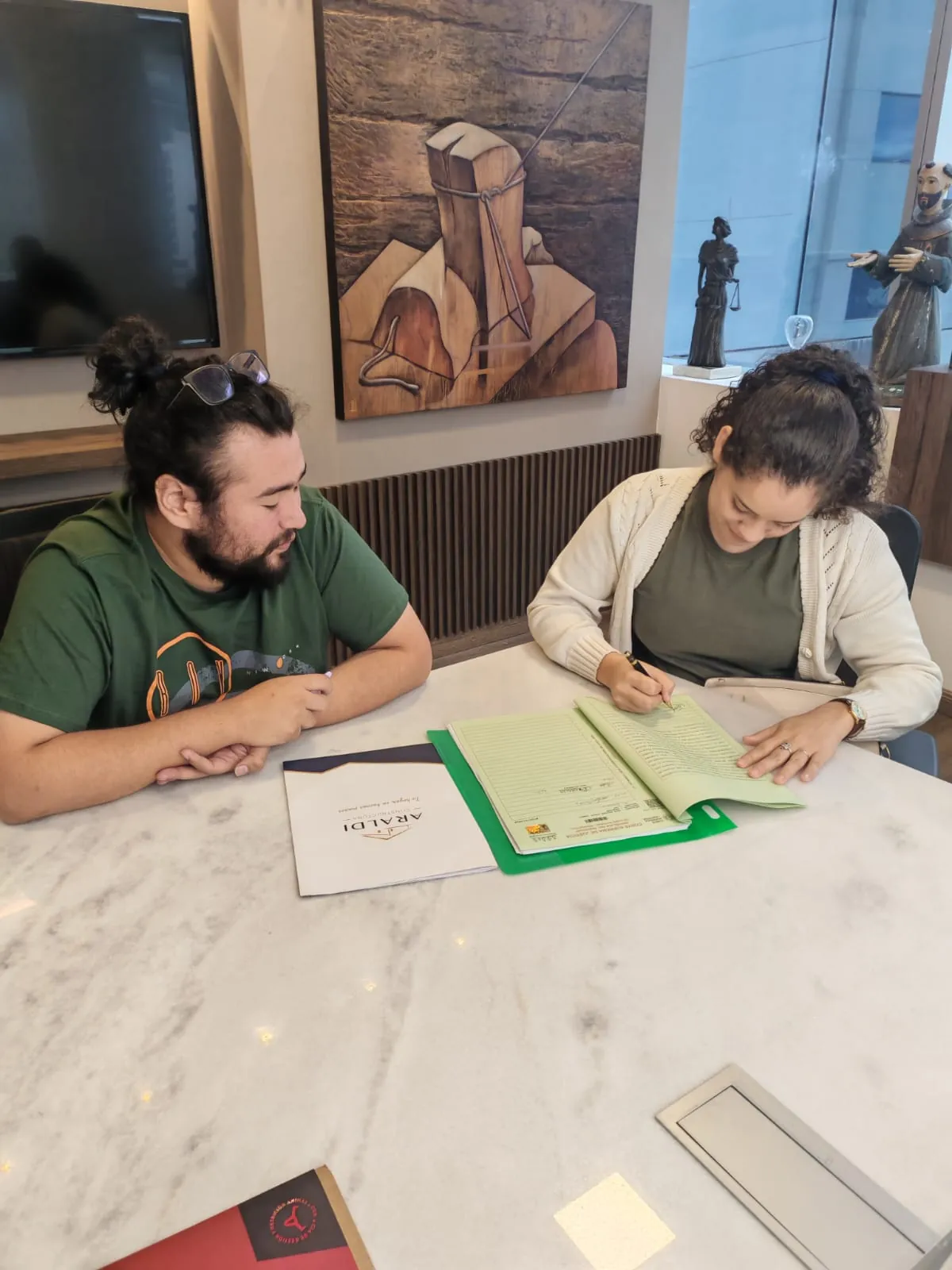 Tami firma documentos en una mesa de mármol mientras Junior espera a su lado. La carpeta de Araldi está abierta.